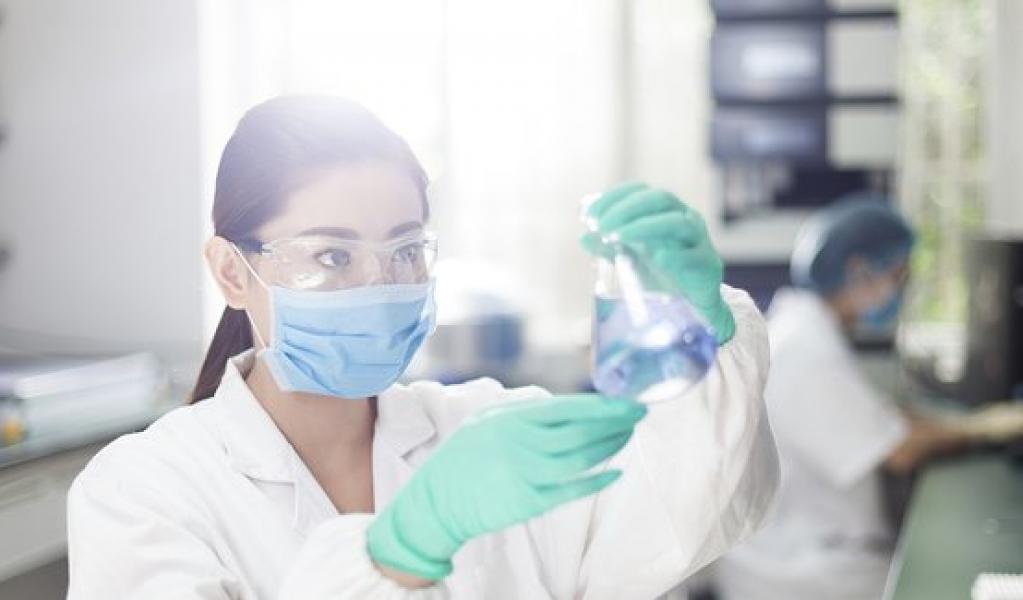 A individual wears a white coat, face mask, and gloves while holding a laboratory beaker.